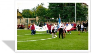 Uroczyste otwarcie boiska orlik. Uczniowe wchodzą na płytę boiska ze sztandarem, za nimi uczniowie niosą flagę Igrzysk olimpijskich. 